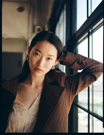 Asian woman poses near window in herringbone blazer and lace top, captured in a medium shot with soft, natural light indoors.