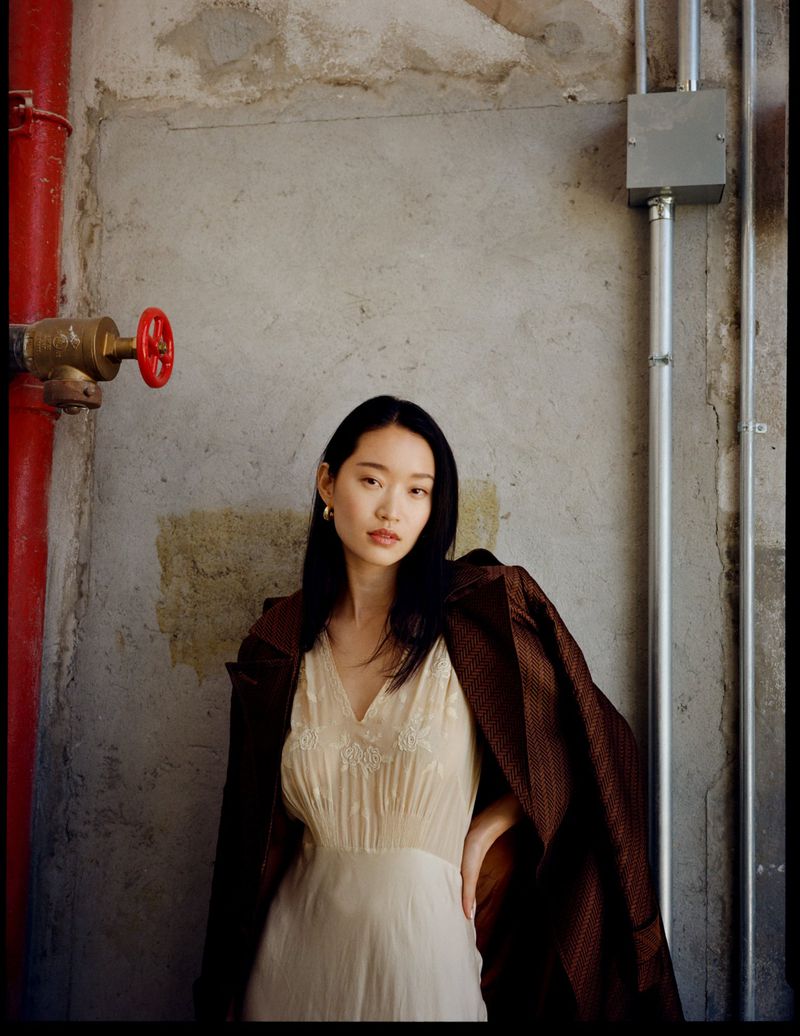 Portrait of an Asian woman in a cream dress and brown jacket, posing against a textured wall with pipes, looking directly at the camera.
