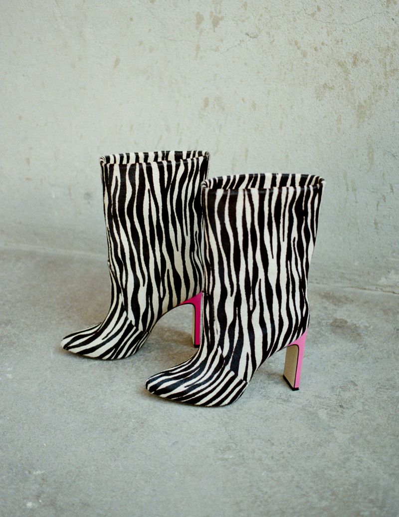 Zebra-striped high-heeled boots with pink block heels on gray surface against gray textured wall background, fashion footwear.