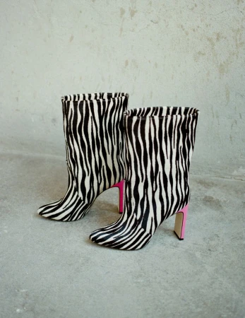 Zebra-striped high-heeled boots with pink block heels on gray surface against gray textured wall background, fashion footwear.