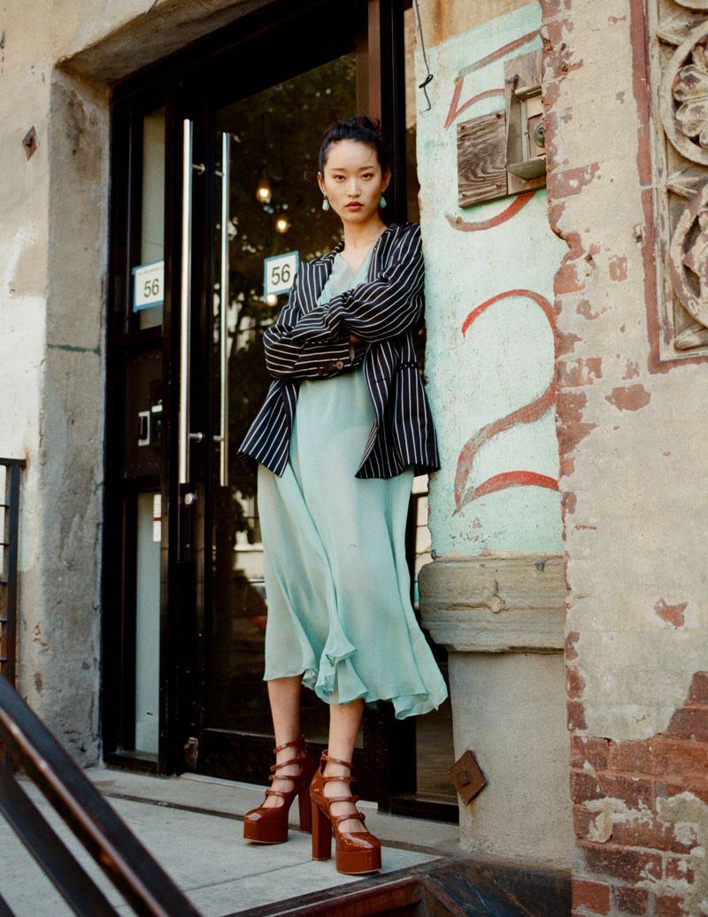 A stylish woman in a striped jacket and light dress poses on stairs next to a weathered building with an interesting architectural element.