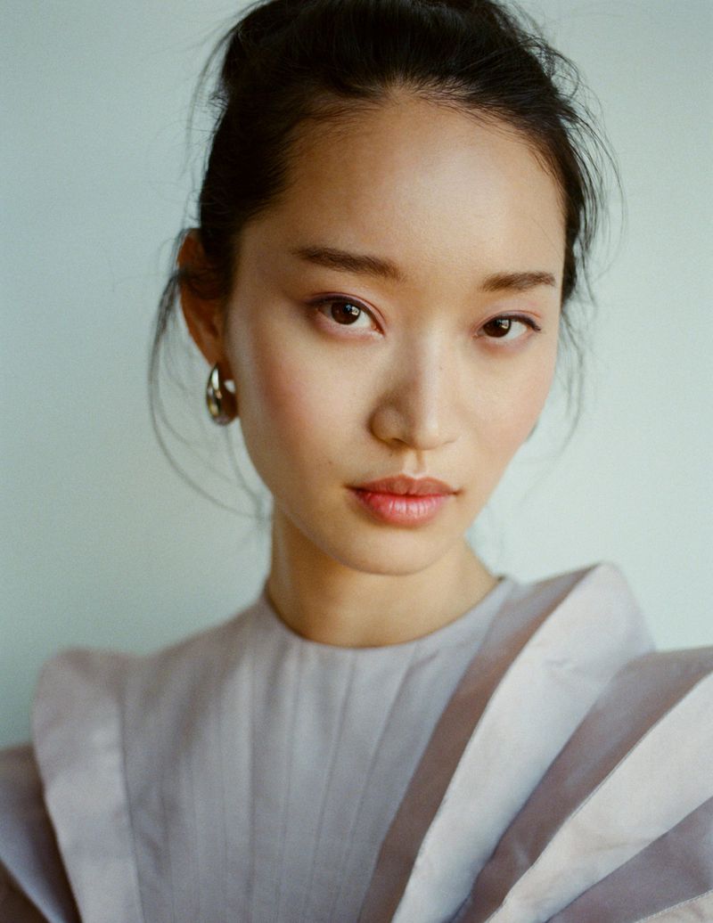 Close-up portrait of an Asian woman with hoop earrings and light purple dress against a plain blueish-grey background in soft, diffused lighting.