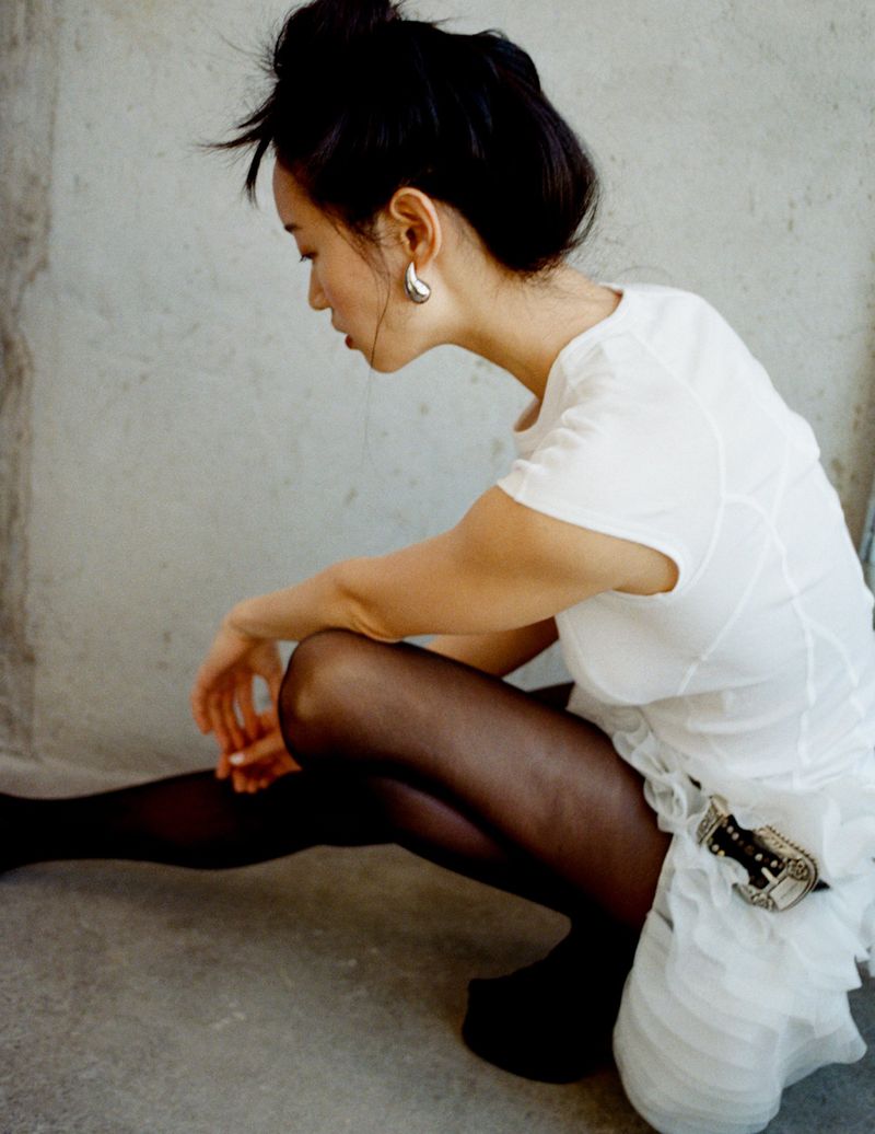 Asian woman in a white dress and black tights poses in a studio against a concrete wall, showcasing fashion-forward style and design elements.