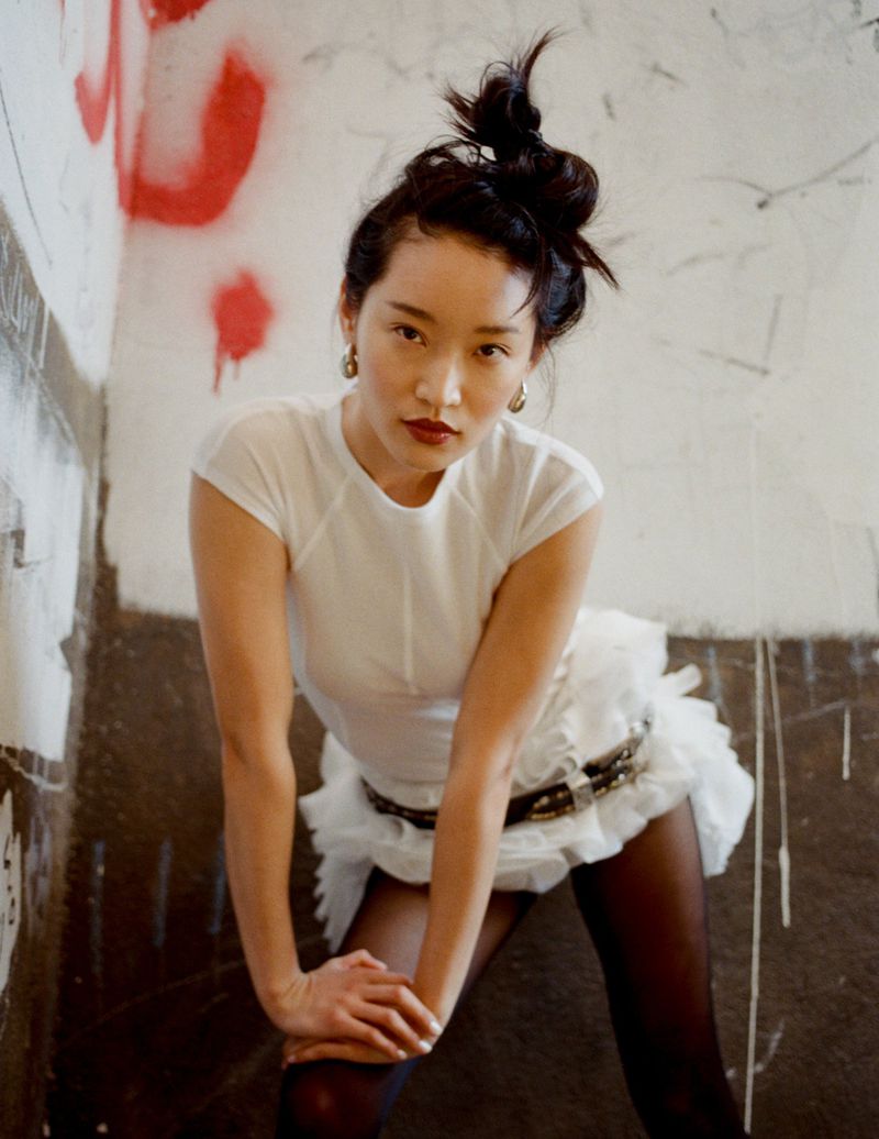 Eye-level shot of an Asian woman in a white outfit with black tights and an elaborate hairstyle, posing in a graffiti-covered corner.