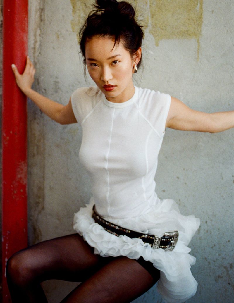 Elegant Asian woman in white dress poses against weathered wall, a bold red pole beside her, blending fashion with urban decay.