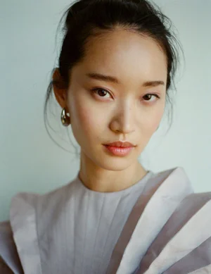 Close-up portrait of an East Asian model with a high bun wearing a grey structured top and silver hoop earrings against a light background.