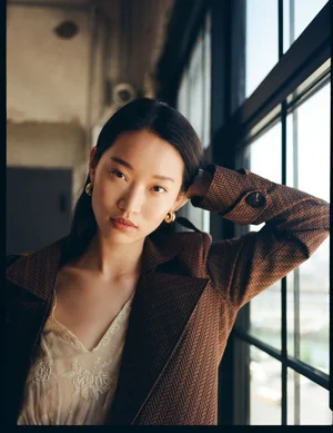 Model in brown herringbone coat and cream lace top posing by a large window for L'Officiel Baltics Revival editorial