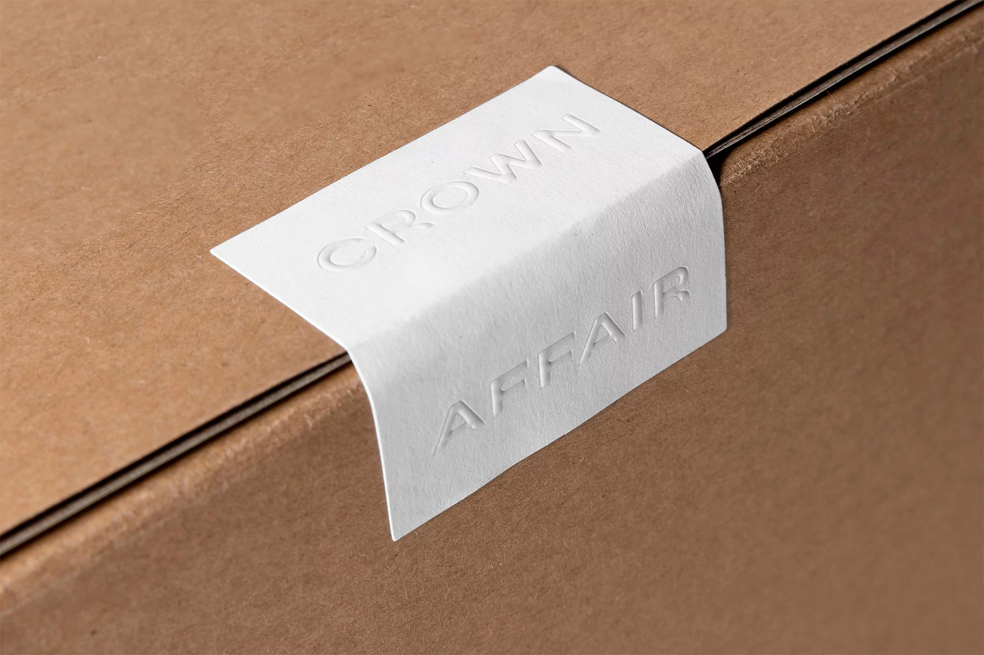 Close-up of a cardboard box with a white embossed label that says 'CROWN AFFAIR', creating a minimalist and textured product presentation.