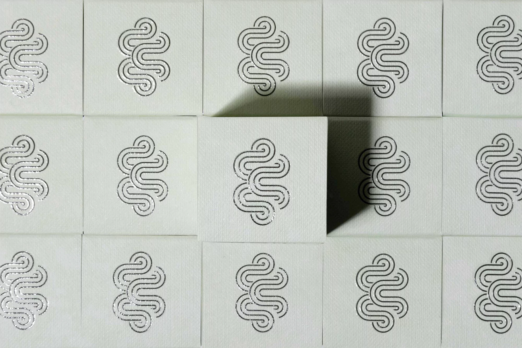 Overhead shot of a grid of square tiles featuring a silver abstract floral design arranged against a white surface with shadows.