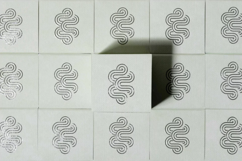 Overhead shot of a grid of square tiles featuring a silver abstract floral design arranged against a white surface with shadows.