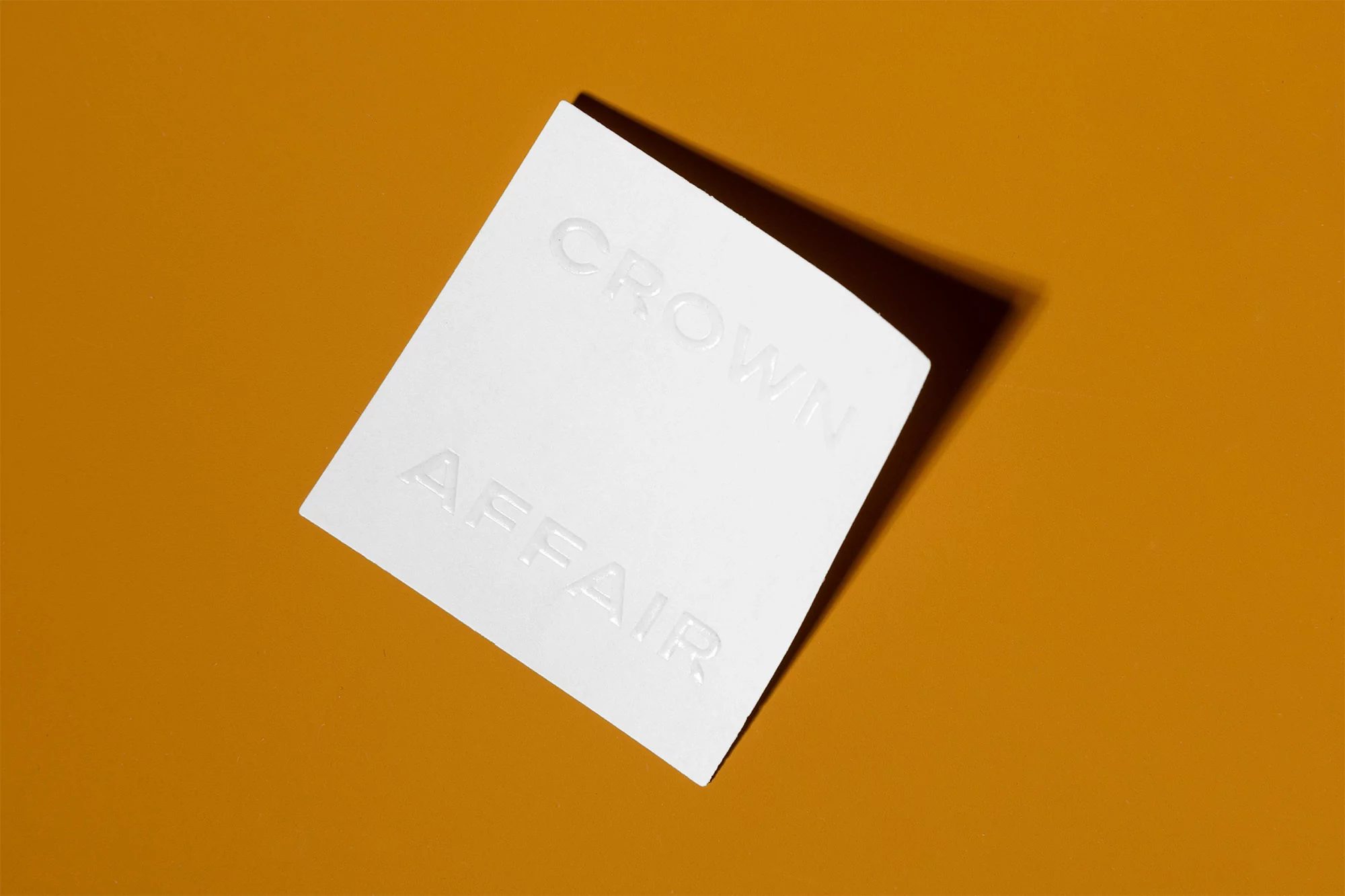 Elevated view of a white invitation or card with 'Crown Affair' text embossed, set against a solid orange background with shadow.