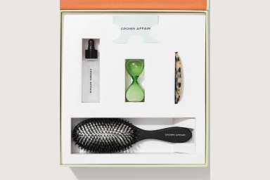 Top-down view of a Crown Affair gift set featuring a hairbrush, serum bottle, green glass hourglass, and a tortoise comb inside a custom white box.