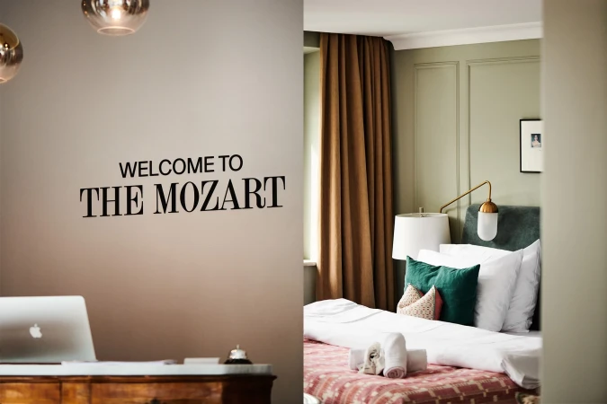 Hotel room interior featuring a bed with pillows and blankets, a desk with a laptop, and wall text 'WELCOME TO THE MOZART'.