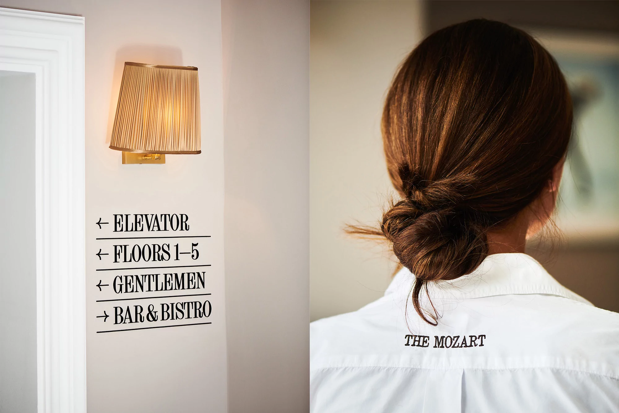 Woman with brunette hair in a bun wears a white collared shirt; a wall has an informational sign and a gold-toned sconce lamp.