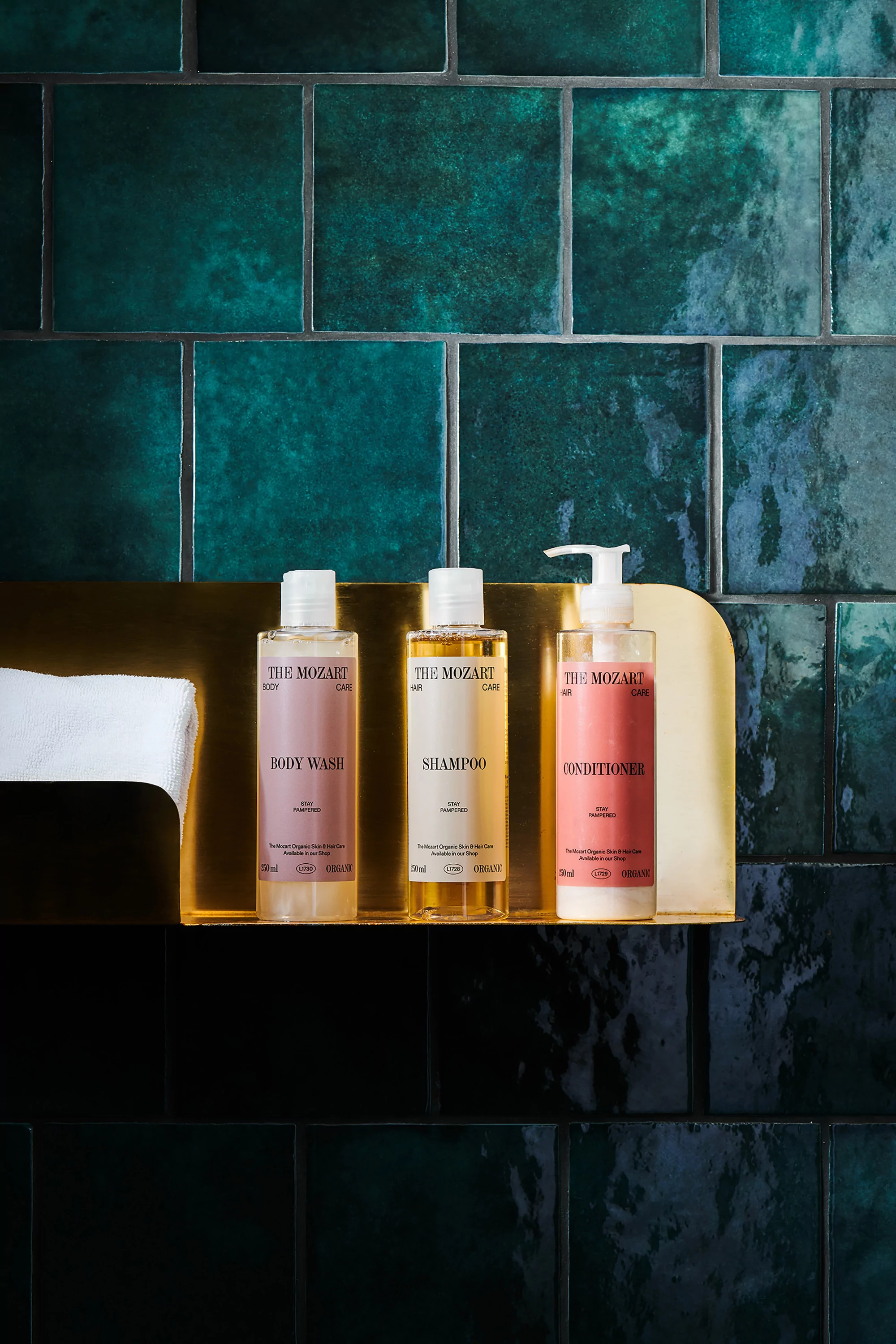 Close-up of a hotel bathroom displaying three labeled bottles of The Mozart body wash, shampoo, and conditioner on a gold shelf against tiled wall.