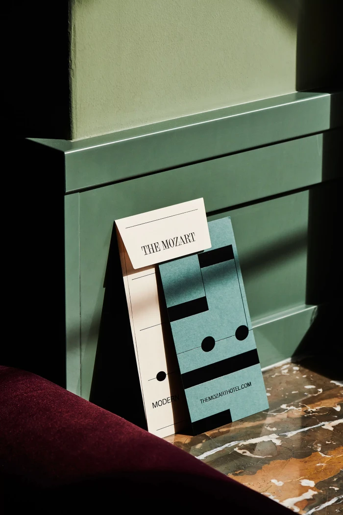 Branded stationery and collateral pieces leaning against a muted green paneled wall with strong natural sunlight casting shadows.