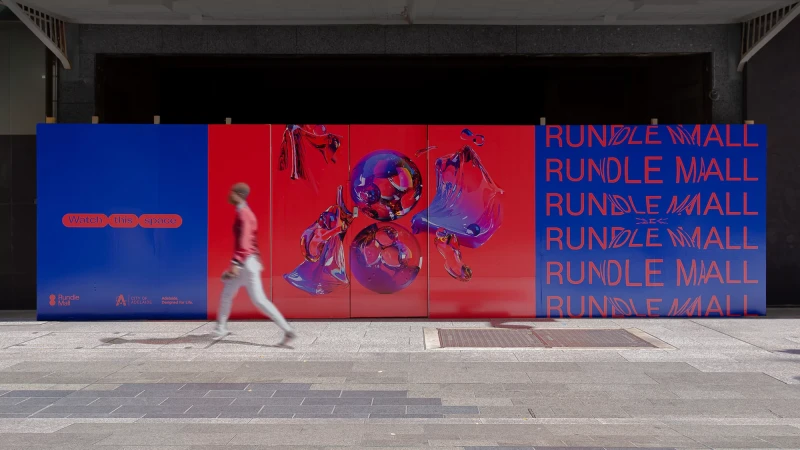Sidewalk view of person walking past Rundle Mall hoarding with abstract art and lettering in red, white, and blue in an urban setting.