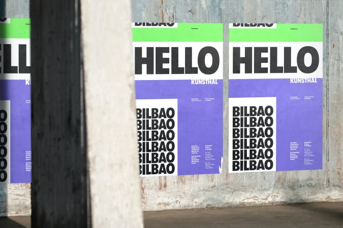 Two promotional posters for a KuntsHal event in Bilbao are adhered to a weathered concrete wall in an urban setting, viewed through a blurred concrete column.