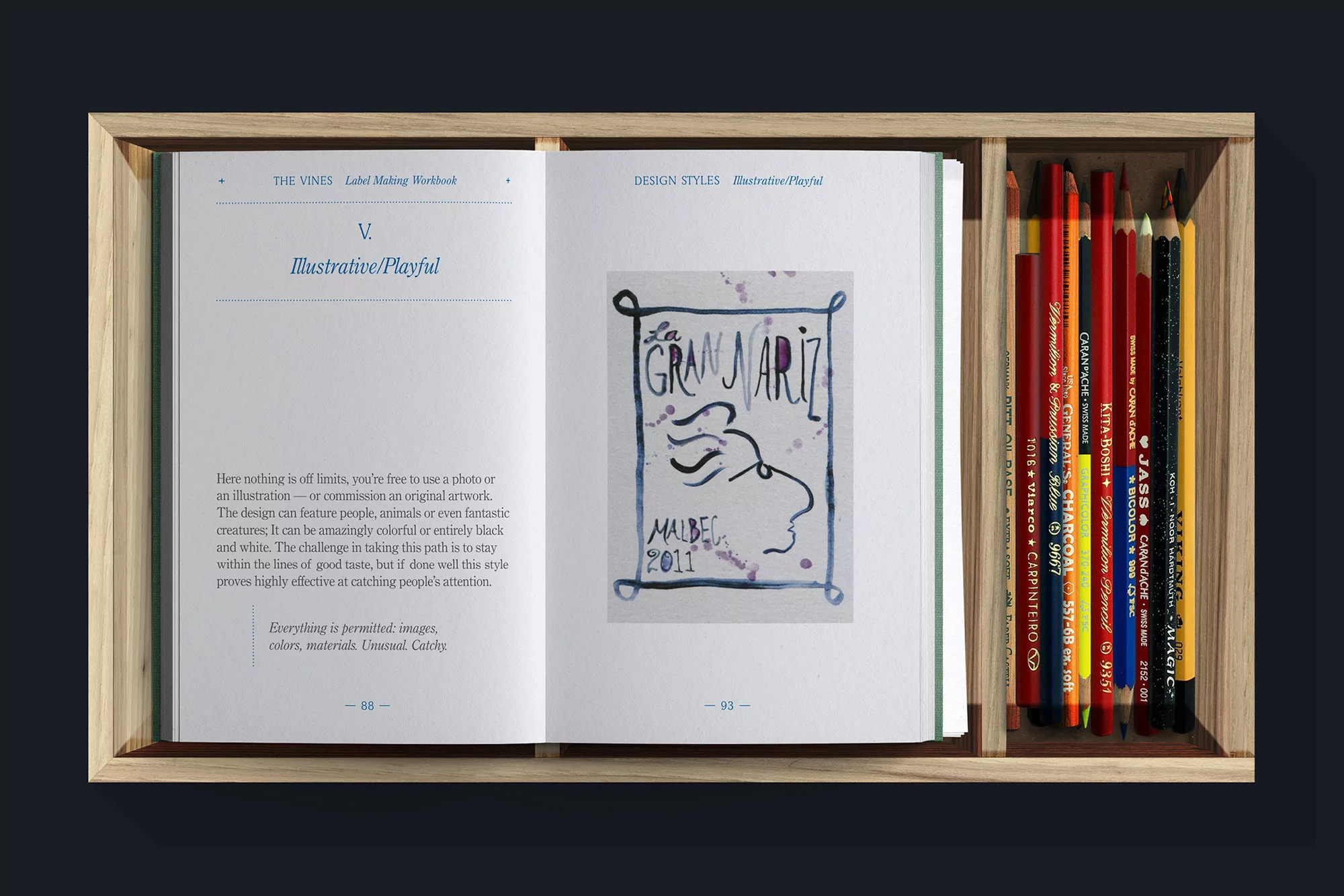 Open book with illustration and text, next to colored pencils. The book displays an illustration of a face and the text 'Gran Nariz'.