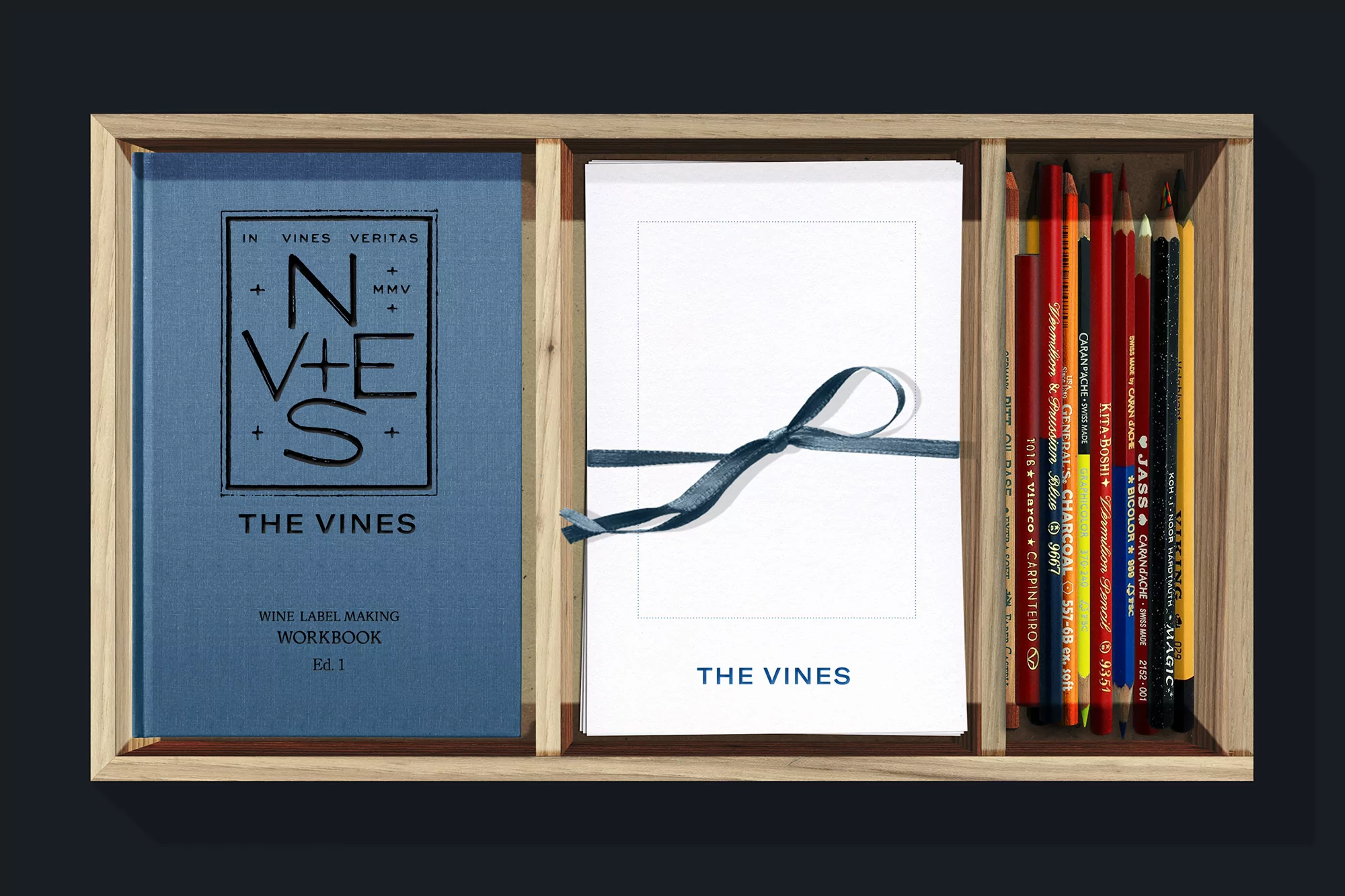 Elevated studio shot of a wooden box set with a blue workbook, white paper, colored pencils, and a blue ribbon.