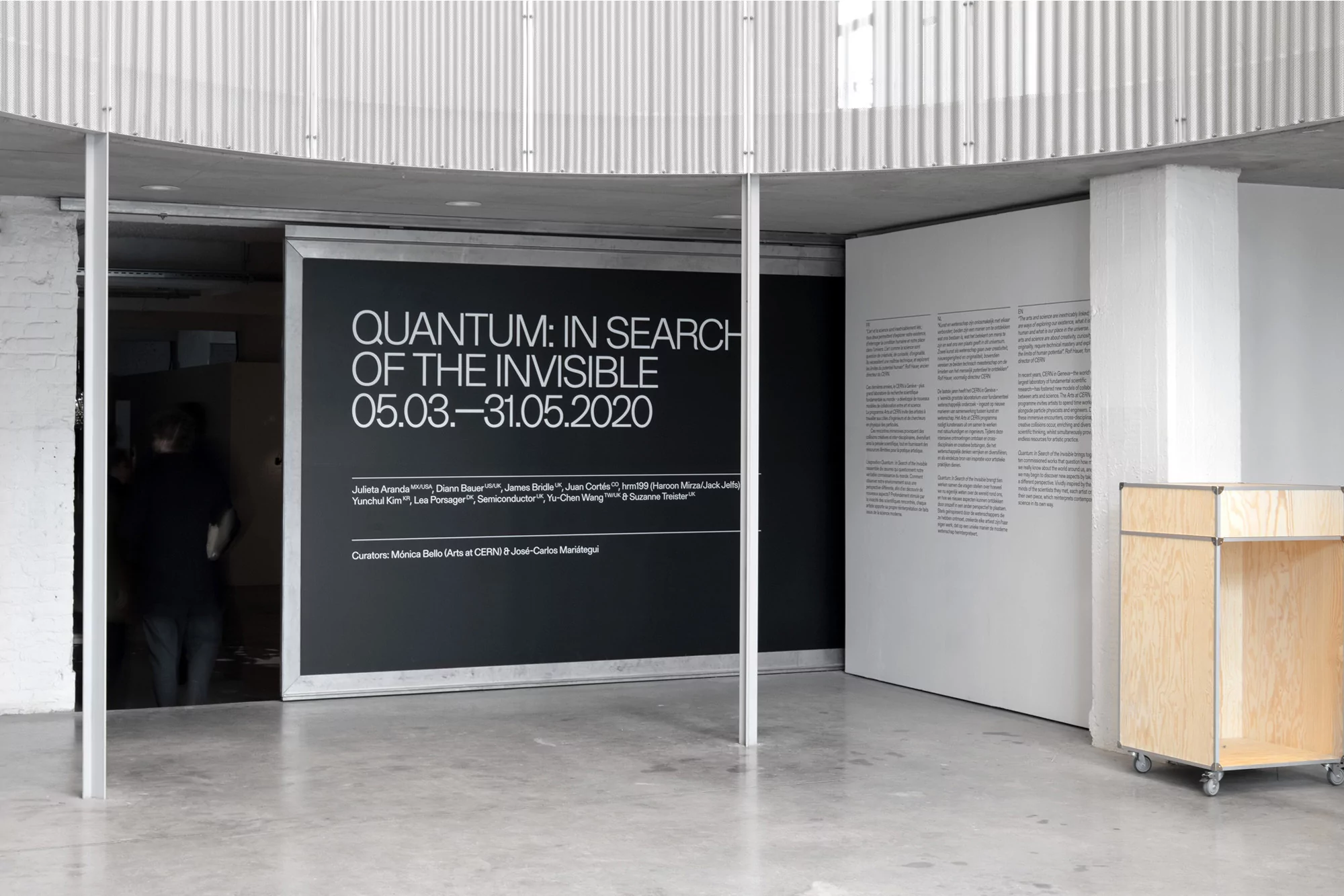 View of an indoor art exhibition with black posters featuring white text, a rolling wooden storage unit, and blurred silhouettes of people.