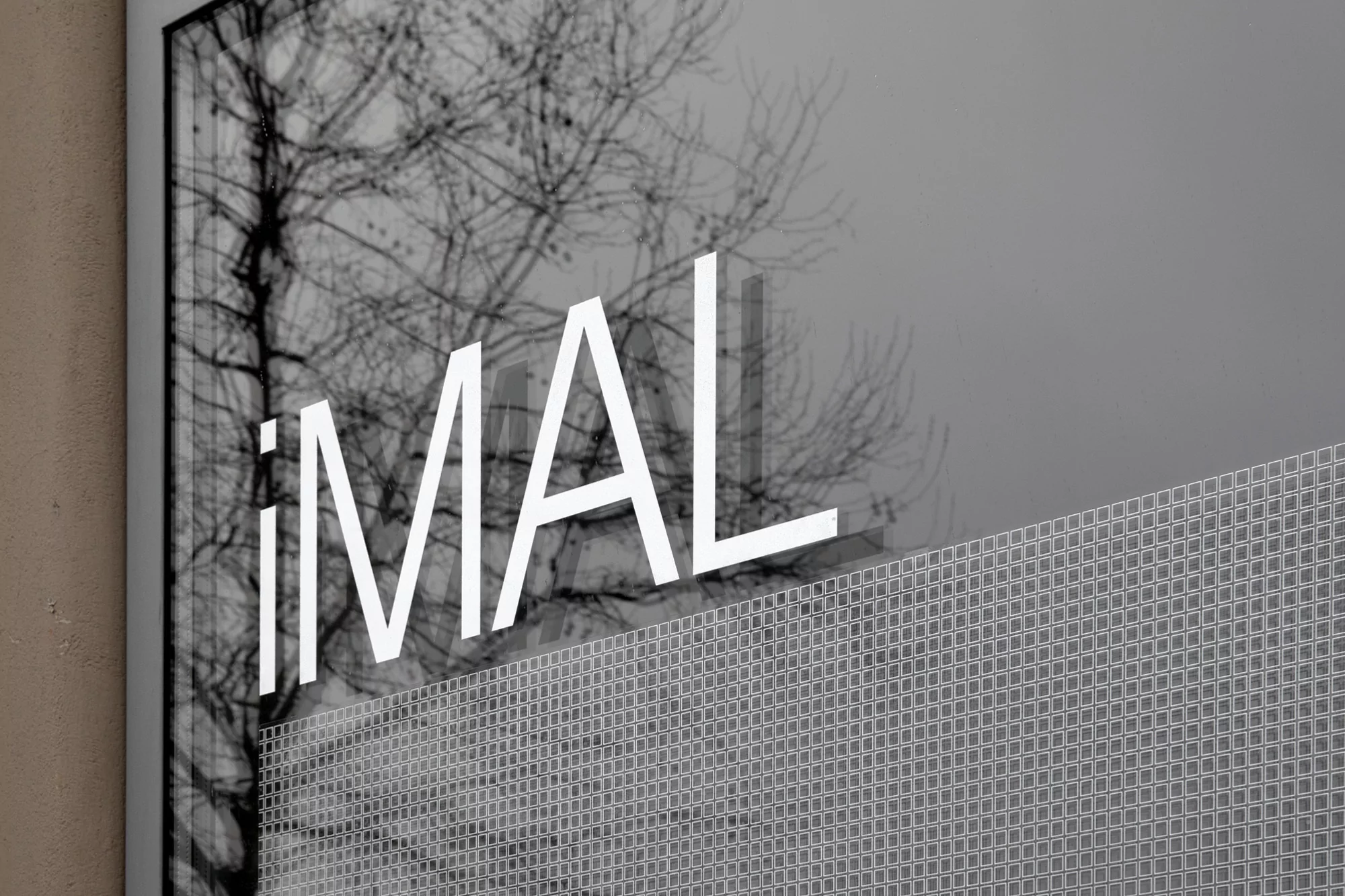 Architectural photograph featuring the text 'iMAL' superimposed on a building exterior, with a bare tree visible in the background.