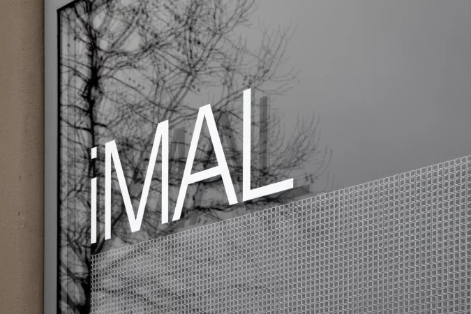 Architectural photograph featuring the text 'iMAL' superimposed on a building exterior, with a bare tree visible in the background.