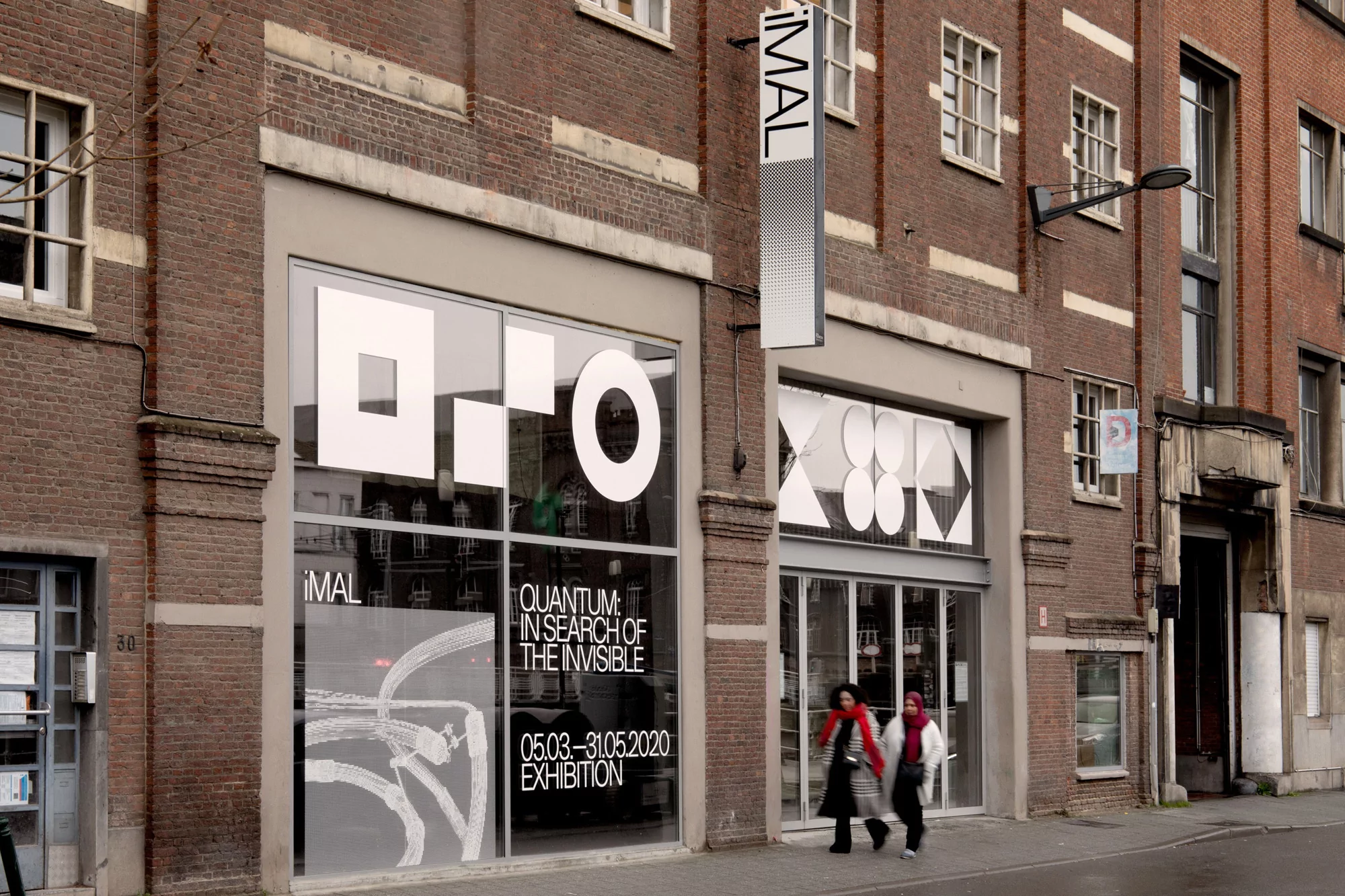 A red brick building with large windows displays geometric art. Two adult females walk by on the street in front of the urban architecture.