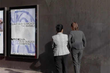 Two women standing on a street next to a backlit exhibition poster for iMAL featuring blue abstract graphics.