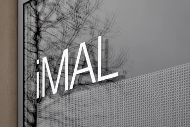 White uppercase text branding reading iMAL on a grey window glass surface reflecting bare tree branches