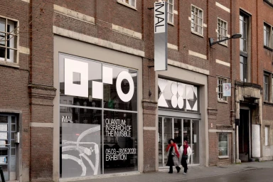 Exterior view of the iMAL building featuring bold white graphic window displays and a vertical hanging sign on a brick street.