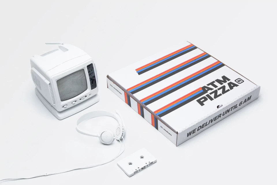 Still life of a white retro TV, headphones, cassette tape, and pizza box with colorful stripes on a white seamless backdrop.