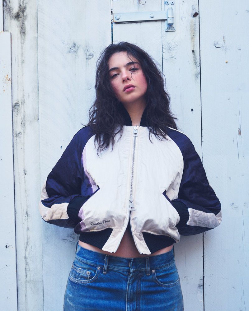 Young woman in a fashion bomber jacket with dark hair poses outdoors against a light grey weathered wooden wall. Casual fashion style.