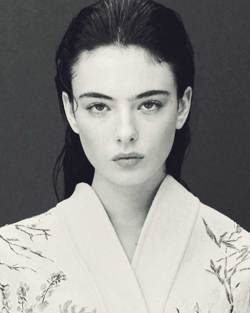 Monochrome portrait of a young woman with dark hair wearing a floral-patterned robe against a dark background, focusing on her serene expression.