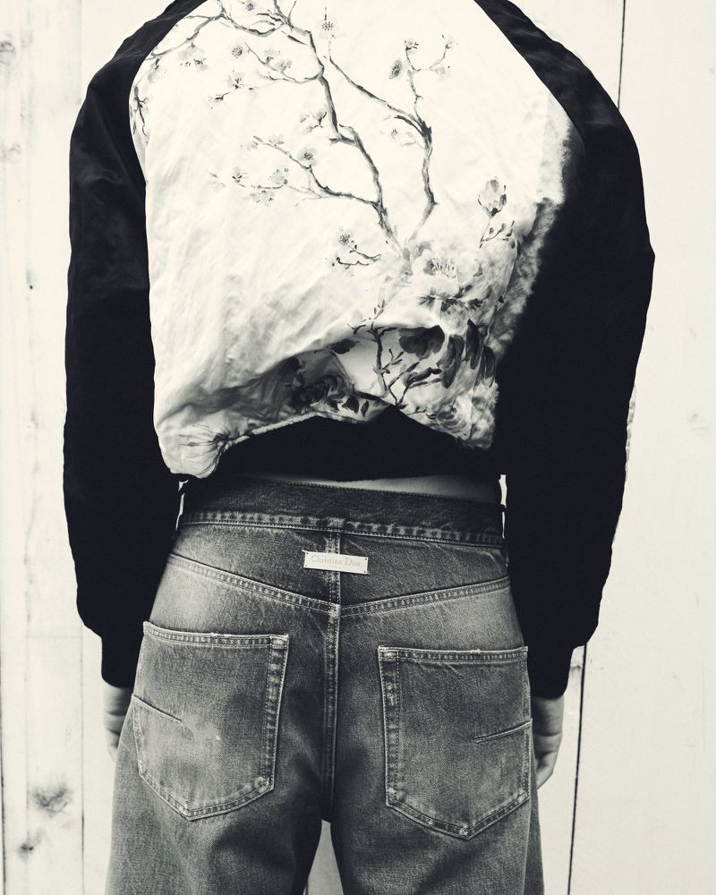 Person wearing a jacket with a tree design and wide-leg denim jeans, posing with their back to the camera in a monochrome setting.