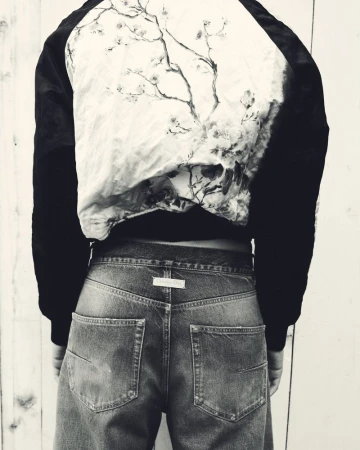 Person wearing a jacket with a tree design and wide-leg denim jeans, posing with their back to the camera in a monochrome setting.