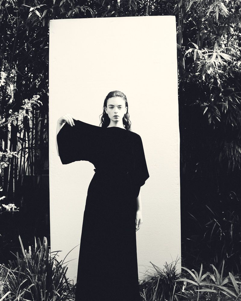 A monochrome fashion shot of a young adult female in an elegant black dress, posing outdoors in front of a white board and leafy background.
