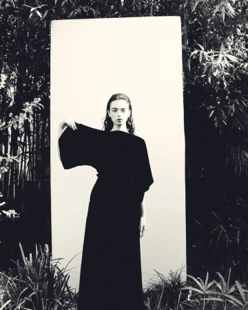 A monochrome fashion shot of a young adult female in an elegant black dress, posing outdoors in front of a white board and leafy background.