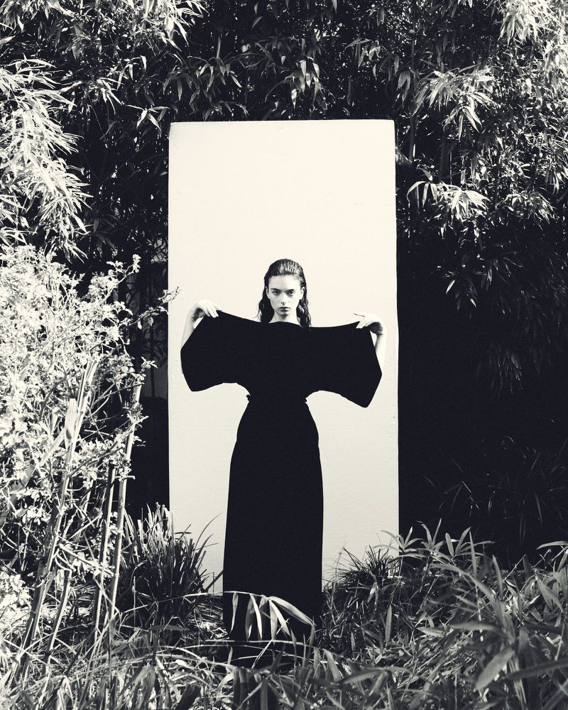 A monochrome fashion shot of a female model in a black dress posing in a lush green outdoor setting with a white screen backdrop.