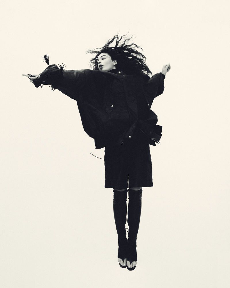 Monochrome fashion shot of a woman with dark hair jumping, wearing a black jacket, skirt, over-the-knee boots against a neutral backdrop.