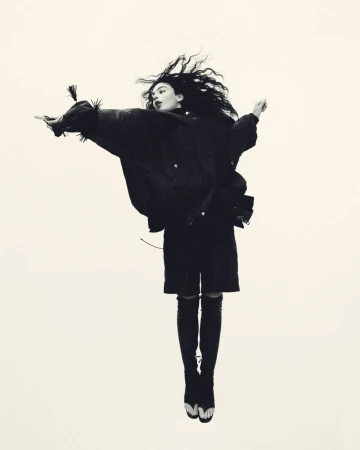 Monochrome fashion shot of a woman with dark hair jumping, wearing a black jacket, skirt, over-the-knee boots against a neutral backdrop.