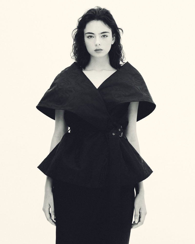 Portrait of an adult female fashion model wearing a dramatic black top with belt detail, set against a clean white studio backdrop.