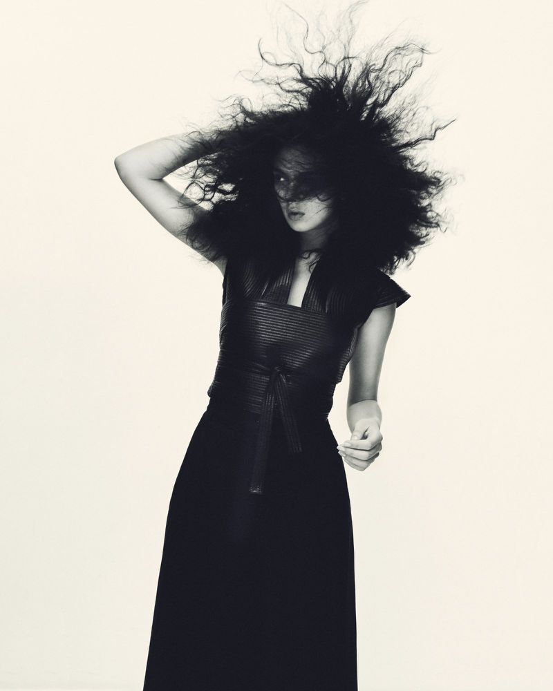 Woman with dramatic dark hair wearing a black dress poses in a studio, captured in a stylish black and white fashion photograph.