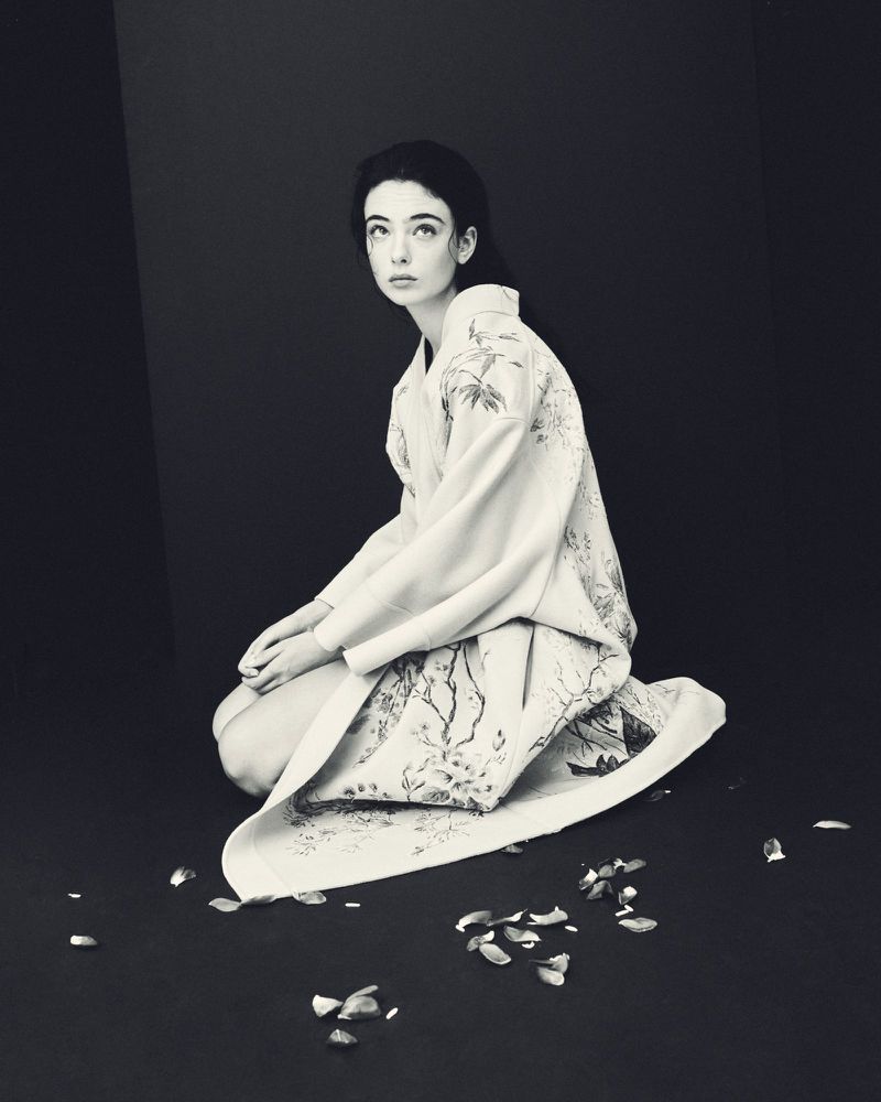 Young woman with fair skin kneels wearing an ornate floral kimono with flower petals scattered around her in a dark studio.