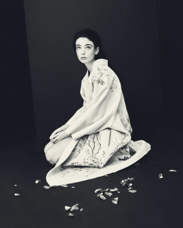 Young woman with fair skin kneels wearing an ornate floral kimono with flower petals scattered around her in a dark studio.