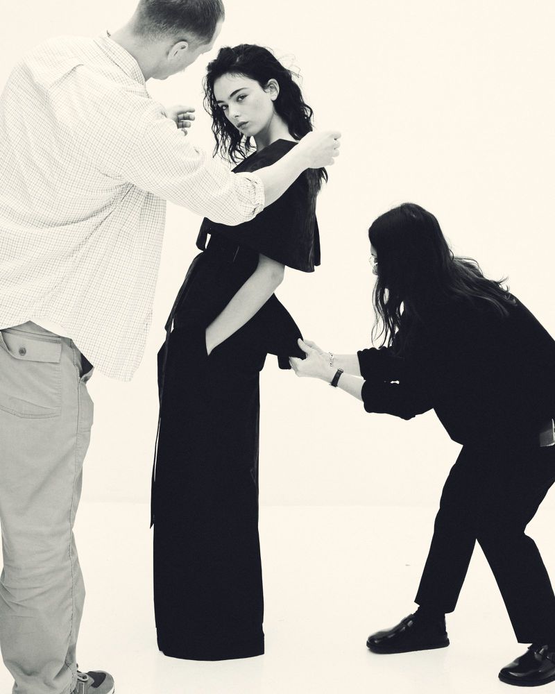 Fashion shoot featuring a model in dark clothing posed between two stylists adjusting her garment in a photo studio with white background.