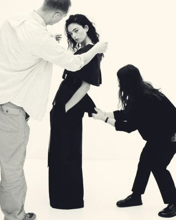 Fashion shoot featuring a model in dark clothing posed between two stylists adjusting her garment in a photo studio with white background.