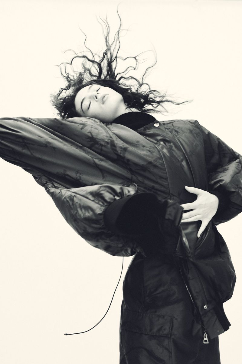 Monochromatic fashion shot of a young adult woman with black, wavy hair, wearing a dark, oversized down jacket against a neutral backdrop.