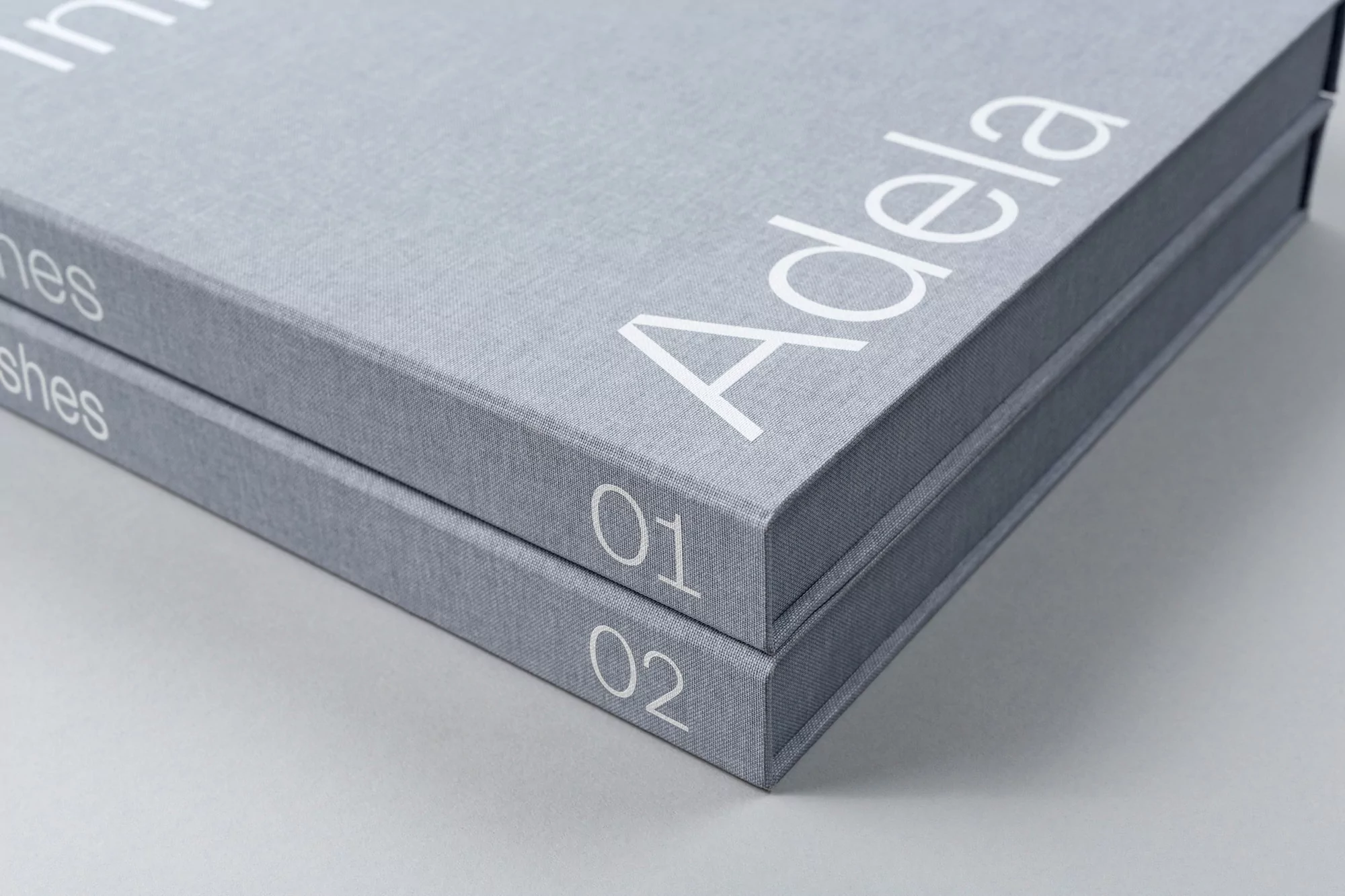 Two grey fabric-bound presentation boxes stacked with white typography reading Adela and numbers 01 and 02