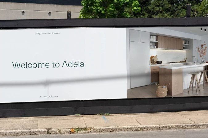 Billboard advertisement featuring a modern kitchen design with white cabinets and a central island, promoting Adela.